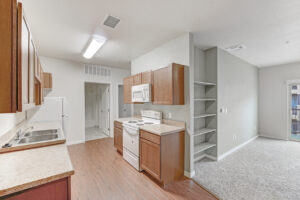 Interior unit kitchen, open floor plan, wood like floors in kitchen area neutral toned wall paint and carpeting, bookshelf on opposite side of stove in living room, laminate countertops, white appliances, light brown cabinetry, dual stainless steel sink, bathroom at end of kitchen hallway.